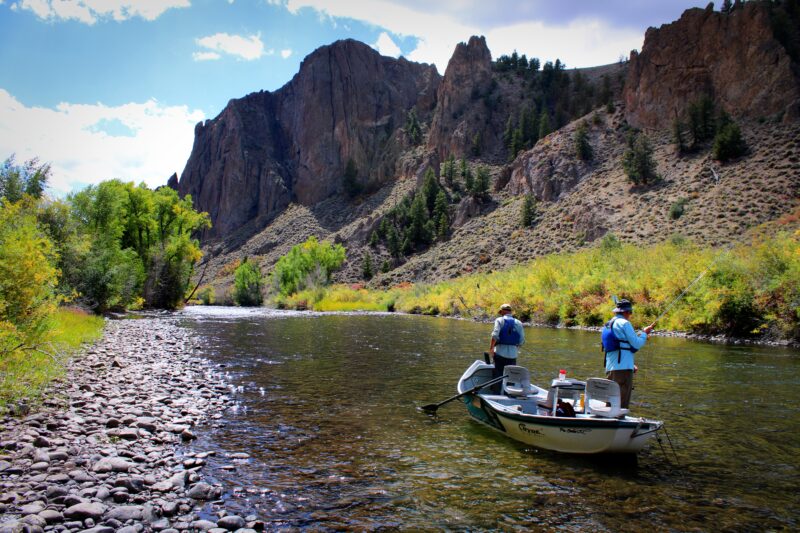Two anglers fly fishing from a boat on a calm river with rocky cliffs in the background.