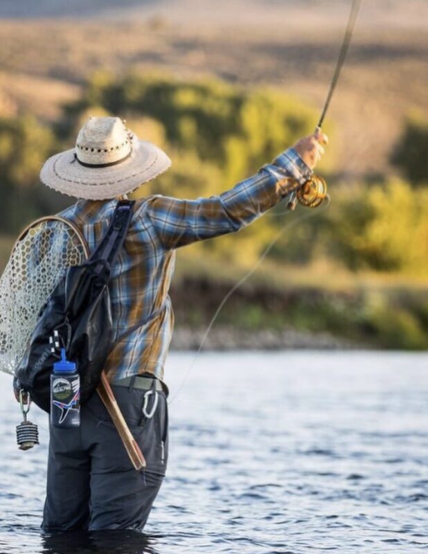 Fly fisherman wearing a straw hat and plaid shirt casting a line in a river.